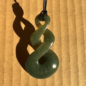 May include: A green jade pendant in a double-twist design, suspended from a black cord. The polished stone reflects light, showcasing its natural variations. The pendant's shadow is cast on a textured surface.