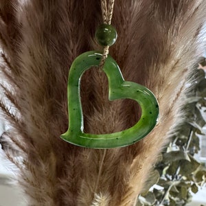 May include: A green jade heart pendant necklace with a brown cord. The heart is carved with a delicate open design.