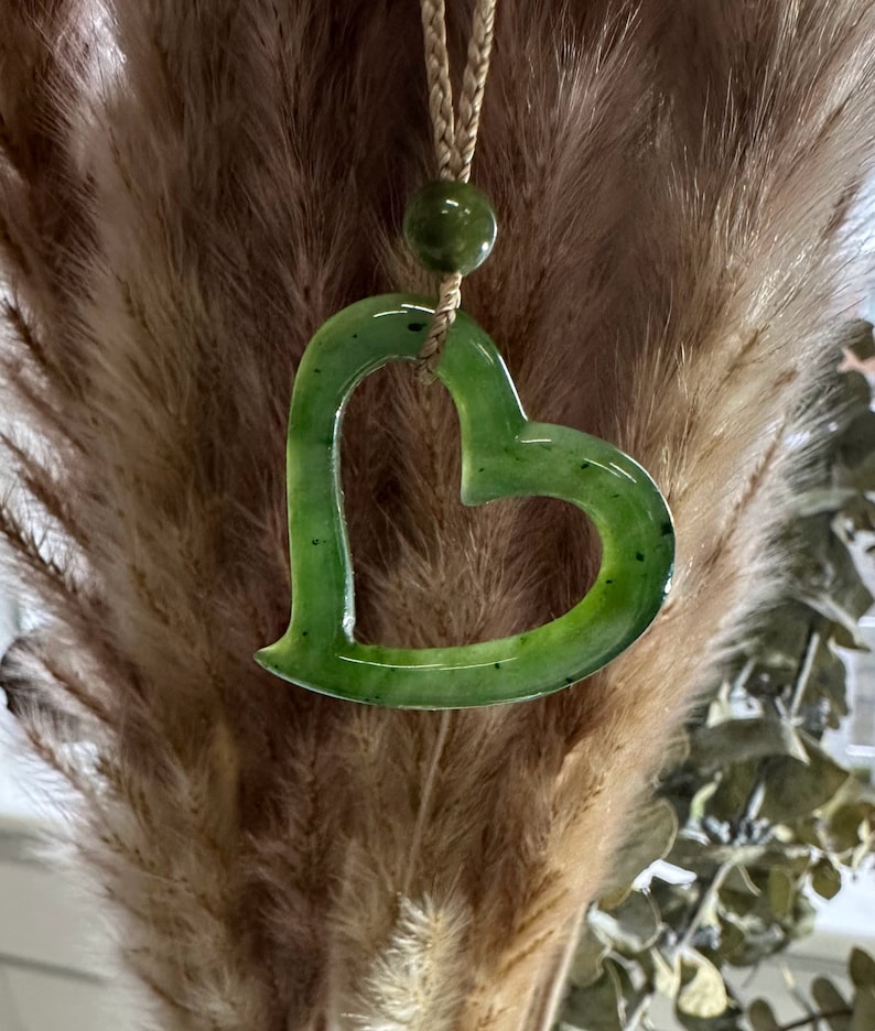 May include: A green jade heart pendant necklace with a brown cord. The heart is carved with a delicate open design.