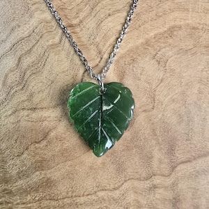 May include: A green jade leaf pendant necklace with a silver-colored chain. The leaf pendant is carved with detailed veining and hangs against a wooden background. The necklace is a nature-inspired accessory.