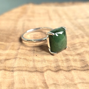 May include: A silver ring with a large, square-cut, deep green gemstone. The gemstone is set in a silver bezel, and the ring is displayed on a wooden surface. The ring's design is simple yet elegant, showcasing the natural beauty of the stone.
