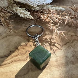 Puede incluir: Un llavero con una piedra preciosa verde en forma de cubo. La piedra está unida a un anillo y una cadena de metal plateado. El fondo presenta flores secas y una superficie de madera.