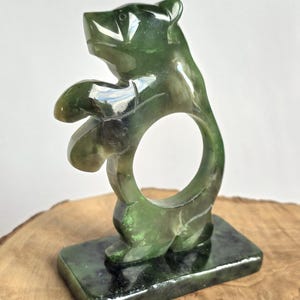 Puede incluir: Una escultura de piedra verde oscuro pulida con la forma de un oso de pie sobre sus patas traseras. El oso tiene una abertura circular en la parte central del cuerpo y está montado sobre una base rectangular del mismo material pétreo moteado.