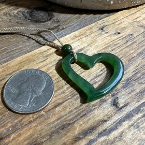 May include: A green jade heart pendant necklace with a simple brown cord. The pendant is a carved heart shape with a hole in the center. The pendant is about the size of a quarter.