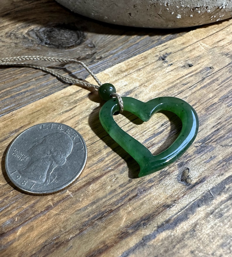 May include: A green jade heart pendant necklace with a simple brown cord. The pendant is a carved heart shape with a hole in the center. The pendant is about the size of a quarter.
