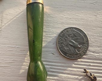 May include: A green, cylindrical object with a silver and gold top, possibly a perfume bottle or a similar container. The object is next to a US quarter coin. The object is approximately 3 inches tall.