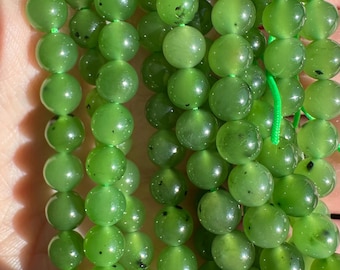 May include: Strands of round, polished green beads. The beads are a vibrant, translucent green color with subtle variations and small dark inclusions. The beads are strung on a green cord, suggesting a necklace or bracelet.