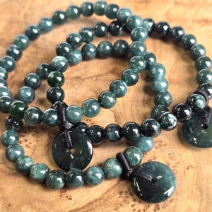 May include: Three jade bead bracelets with dark green beads and round, flat jade pendants. The bracelets are on a wooden surface. Each bracelet has a black cord attaching the pendant.