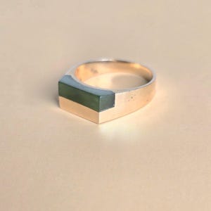 May include: A gold ring featuring a rectangular, dark green stone inlay set into the top of the band. The band has a smooth, polished finish and a geometric shape, with the green stone flush against the metal surface of the ring.