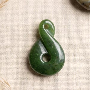 May include: A polished, green jade pendant in a figure-eight shape. The pendant has two circular openings and a smooth, glossy surface. The jade has natural variations in color, with darker green spots.