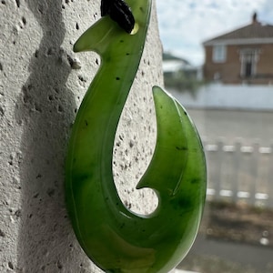 Puede incluir: Un colgante de jade verde en forma de gancho, suspendido por un cordón negro. La piedra pulida tiene variaciones naturales de color y está colocada contra una pared texturizada de color claro. El colgante es un diseño maorí tradicional.