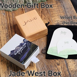 May include: A wooden gift box with the word "JADE" engraved on the lid. The box is sitting on a wooden surface next to a white box with a mountain scene and the text "West Coast Jade by Jade West". There is also a black velvet bag with two white cards that say "Jade Jewelry" on them.