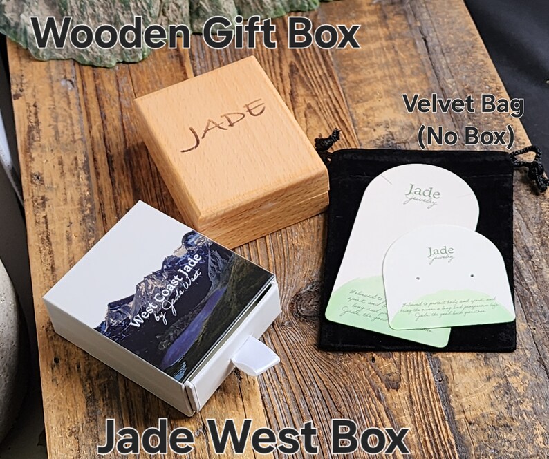 May include: A wooden gift box with the word "JADE" engraved on the lid. The box is sitting on a wooden surface next to a white box with a mountain scene and the text "West Coast Jade by Jade West". There is also a black velvet bag with two white cards that say "Jade Jewelry" on them.