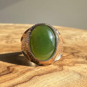 May include: A ring featuring a large, oval-shaped, smooth green stone set in a textured, metallic band. The metal has a rough, irregular surface finish that contrasts with the polished, glossy appearance of the central green gemstone.