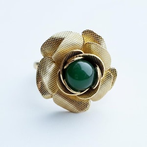 May include: A gold-colored ring featuring a textured rose design. The center of the rose holds a round, dark green gemstone. The ring is set against a white background, highlighting the intricate details of the floral design.