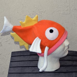 May include: An orange fish-shaped hat with yellow fins and a white tail. The hat has a pink band around the base and a large white eye with a black pupil. A fun, whimsical costume accessory.