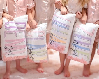 May include: Five young people wearing matching light pink pajama sets, each holding a white pillow with a name printed on it in a pastel color scheme. The pillows have a decorative pink filler at the bottom.