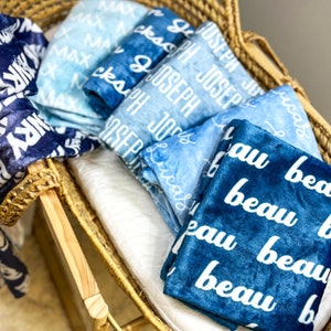 May include: A wicker basket filled with blue and white personalized blankets. The blankets have names written on them, including "Max", "Joseph", and "Beau".
