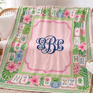 May include: A soft pink throw blanket featuring a central dark blue monogram with the letters SBL. The border displays a repeating green geometric pattern, watercolor illustrations of mahjong tiles, pink hibiscus flowers, and blue pottery.
