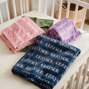 May include: A collection of four soft blankets in navy blue, pink, purple, and olive green. Each blanket features a repeating white script pattern of a specific name: Grayson, Harper, Sophia, or Cooper, printed across the fabric surface.