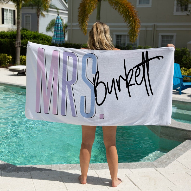 May include: White beach towel with the text "MRS. Burkett" in pink, blue, and black lettering.