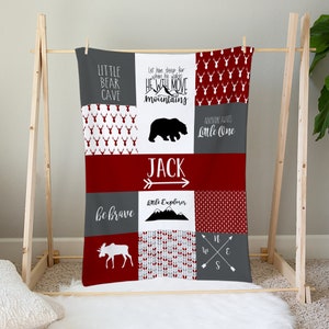 May include: A red and gray baby blanket with a patchwork design. The blanket features a variety of woodland animals, including a bear, moose, and deer. The blanket also includes the text "Little Bear Cave", "Let him sleep for when he wakes he will move mountains", "Adventure awaits Little One", "Jack", "Little Explorer", "Be brave", and "N W E S".