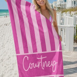 May include: A pink and light pink striped beach towel with the name "Courtney" in white script. The towel is held up on a sunny day at the beach.