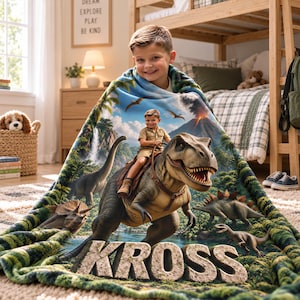 May include: A soft fleece blanket featuring a vibrant prehistoric jungle scene with a large T-Rex, various dinosaurs, a volcano, and a child riding the T-Rex. The name KROSS is printed in large, textured, stone-like letters at the bottom.