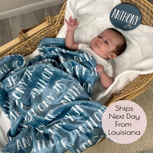 May include: A blue baby blanket with the name "Anthony Jr." printed in white on it. The blanket is folded and placed in a wicker basket with a baby lying on top of it. A round wooden sign with the name "Anthony Jr." is placed on the side of the basket.