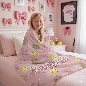 May include: A pink softball-themed blanket with a personalized name. The blanket features yellow softballs, brown gloves, and pink bows. The name "Kayleigh" is written in a cursive font. The blanket is on a bed in a pink room.