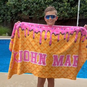 May include: A large, rectangular beach towel with a waffle cone design. The top of the towel has a pink frosting design with brown drips and sprinkles. The towel has the name "JOHN MAR" printed in large, pink letters.