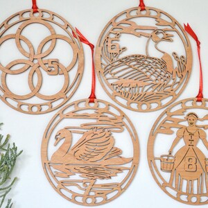 May include: Four wooden Christmas ornaments with red ribbons. Each ornament is circular with intricate cut-out designs. The ornaments depict the numbers 5, 6, 7, and 8, along with corresponding images of a swan, geese, and a woman with buckets.