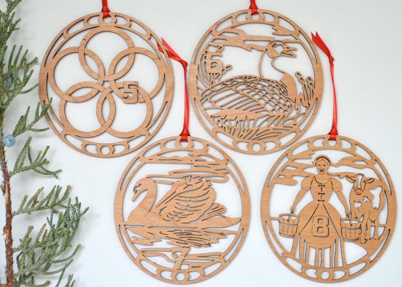 May include: Four wooden Christmas ornaments with red ribbons. Each ornament is circular with intricate cut-out designs. The ornaments depict the numbers 5, 6, 7, and 8, along with corresponding images of a swan, geese, and a woman with buckets.