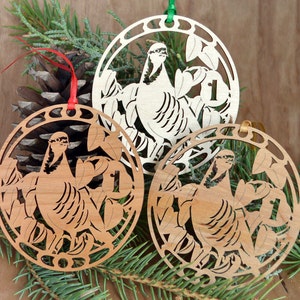 May include: Three wooden Christmas ornaments with a bird design. Each ornament is round with a laser-cut bird and leaf design. The number "1" is visible on each ornament. The ornaments are made of light and medium-toned wood and have red, green, and gold ribbons.