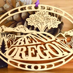 May include: Oval wooden ornament with a laser-cut design featuring the text "BEND OREGON" over a mountain landscape with trees. The ornament has a purple ribbon for hanging.