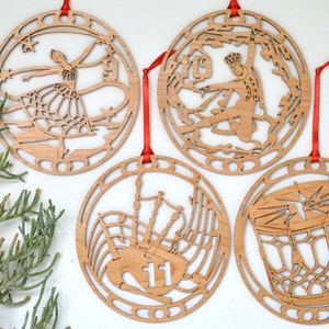 May include: Four wooden Christmas ornaments with red ribbon hangers. Each ornament features a different cutout design: a ballerina, a figure skater, bagpipes with the number 11, and a drum. The ornaments are made of light-colored wood.