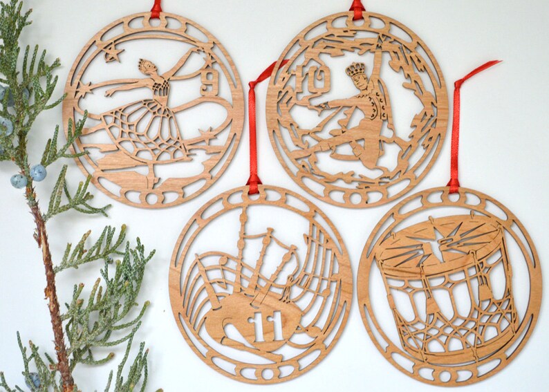 May include: Four wooden Christmas ornaments with red ribbon hangers. Each ornament features a different cutout design: a ballerina, a figure skater, bagpipes with the number 11, and a drum. The ornaments are made of light-colored wood.