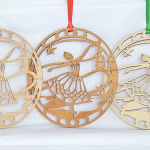 May include: Three wooden ballerina ornaments with different colored ribbons. Each ornament features a cutout design of a ballerina in a circular frame with stars and architectural elements. The ribbons are gold, red, and green.