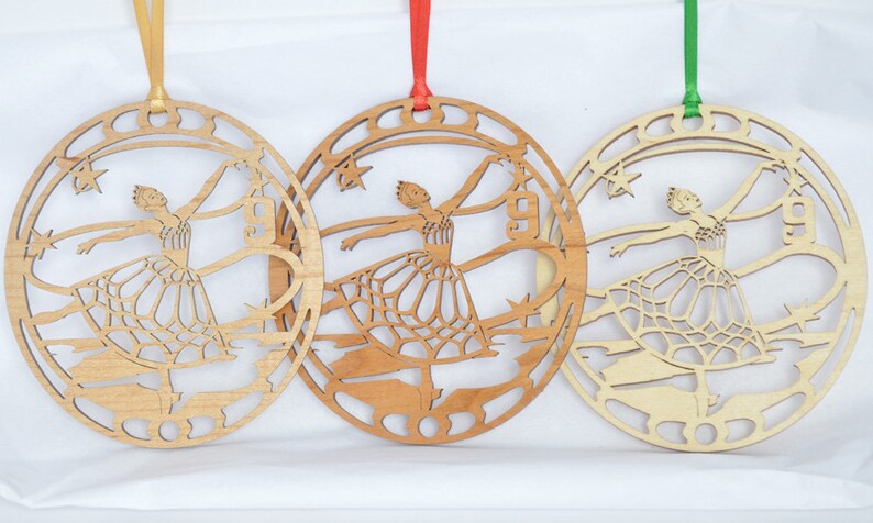 May include: Three wooden ballerina ornaments with different colored ribbons. Each ornament features a cutout design of a ballerina in a circular frame with stars and architectural elements. The ribbons are gold, red, and green.