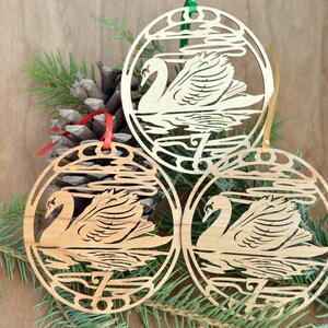 May include: Three wooden swan ornaments with red, green, and gold ribbons for hanging. Each ornament features a swan design within a circular frame, with water and cloud details. The ornaments are displayed with pine needles and a pine cone.