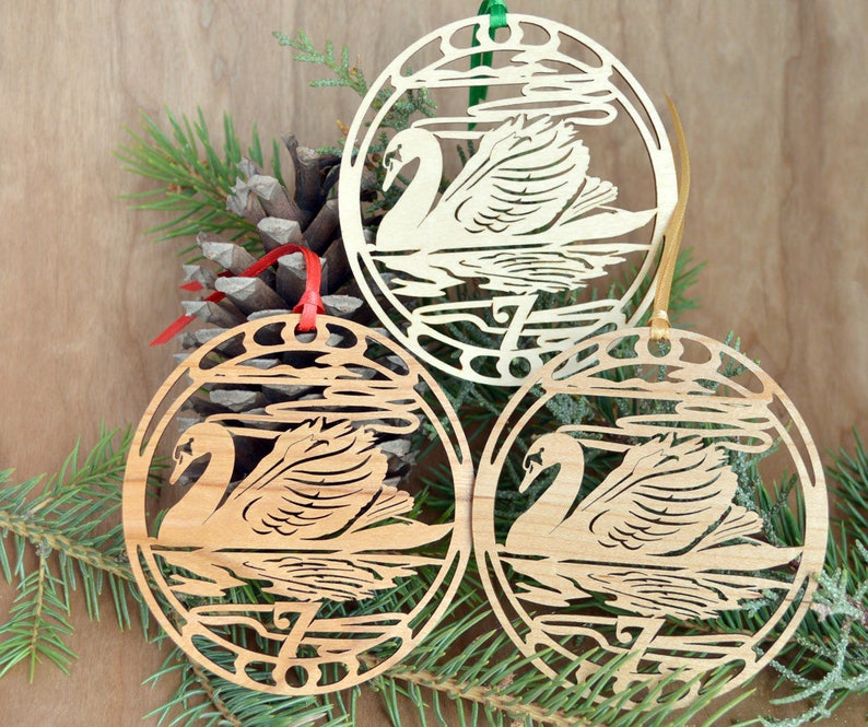 May include: Three wooden swan ornaments with red, green, and gold ribbons for hanging. Each ornament features a swan design within a circular frame, with water and cloud details. The ornaments are displayed with pine needles and a pine cone.