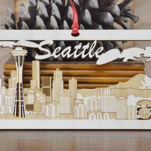 May include: Wooden rectangular ornament featuring a laser-cut design of the Seattle skyline, including the Space Needle and the city name. The ornament has a red ribbon for hanging and is made of light-colored wood.