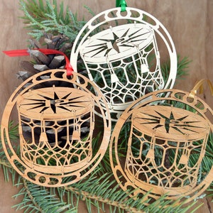 May include: Three wooden Christmas ornaments with a drum design. The ornaments are laser-cut with intricate details, featuring a drum shape with a star in the center. Each ornament has a different color ribbon for hanging. The ornaments are displayed with pine needles and a pine cone.