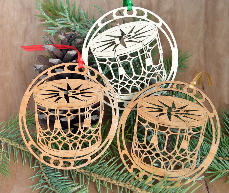 May include: Three wooden Christmas ornaments with a drum design. The ornaments are laser-cut with intricate details, featuring a drum shape with a star in the center. Each ornament has a different color ribbon for hanging. The ornaments are displayed with pine needles and a pine cone.