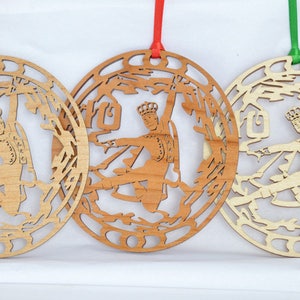 May include: Three wooden ornaments with a cutout design of a figure in a crown, possibly a king or prince, within a circular frame. Each ornament has a different wood tone and a ribbon for hanging. The ribbons are gold, red, and green.