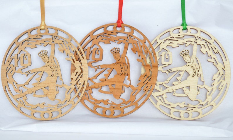 May include: Three wooden ornaments with a cutout design of a figure in a crown, possibly a king or prince, within a circular frame. Each ornament has a different wood tone and a ribbon for hanging. The ribbons are gold, red, and green.