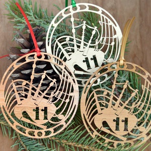 May include: Three wooden Christmas ornaments with a bagpipe design and the number 11. The ornaments are circular with a cut-out design of musical notes and a bagpipe. Each ornament has a different colored ribbon for hanging.