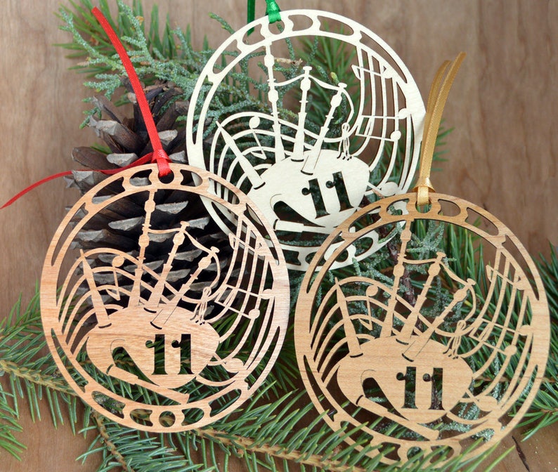 May include: Three wooden Christmas ornaments with a bagpipe design and the number 11. The ornaments are circular with a cut-out design of musical notes and a bagpipe. Each ornament has a different colored ribbon for hanging.