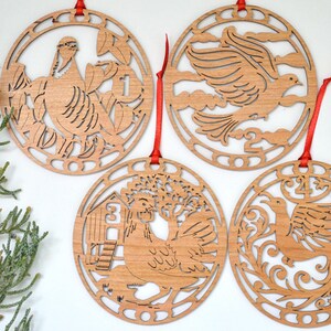 May include: Four wooden Christmas ornaments with red ribbons. Each ornament features a different laser-cut design: a partridge in a pear tree, a dove, a hen with a coop, and a phoenix. The ornaments are circular and made of light-colored wood.
