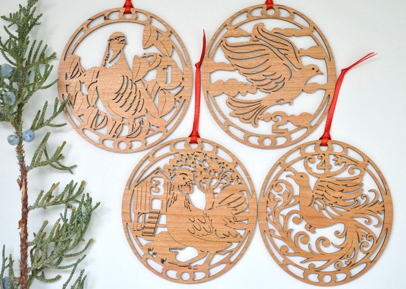 May include: Four wooden Christmas ornaments with red ribbons. Each ornament features a different laser-cut design: a partridge in a pear tree, a dove, a hen with a coop, and a phoenix. The ornaments are circular and made of light-colored wood.